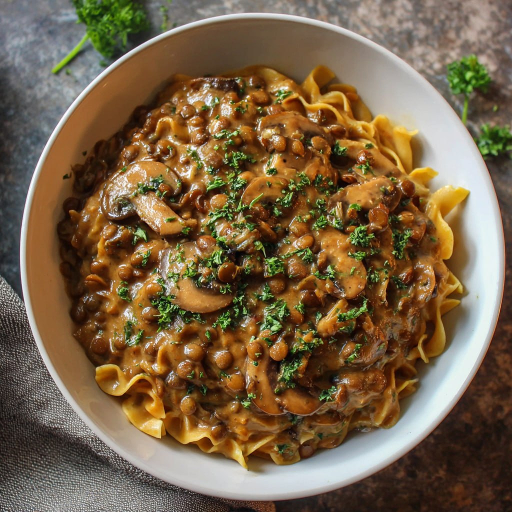 Dairy-free mushroom lentil stroganoff plated with noodles garnished with parsley and a drizzle of olive oil