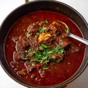 Bowl of Texas chili with chunks of beef, deep red broth, melted cheese, sour cream, jalapeños, and cornbread