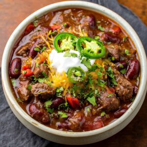 Bowl of spicy steak chili with chunks of seared beef, beans, peppers, melted cheese, sour cream, and cilantro