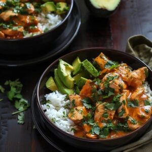 Chicken rice bowl with avocado on a wooden board, golden seared chicken cubes over fluffy white rice, half a sliced ripe avocado fanned across the top, fresh cilantro, lime wedges, drizzle of chipotle crema