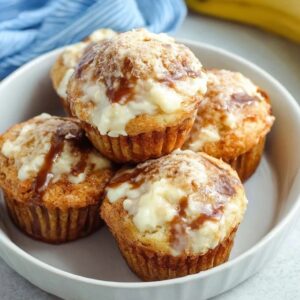 Banana cream cheese muffins on a wooden board, golden banana muffins with visible white cream cheese centers, sliced banana slices on top, sprinkle of brown sugar